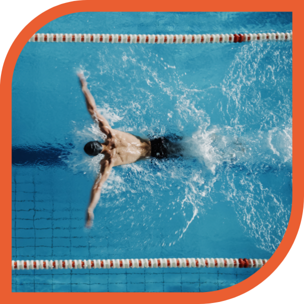 A imagem mostra uma pessoa nadando em uma piscina. A foto é tirada de cima, capturando a pessoa no meio de uma braçada de nado borboleta, com os braços estendidos para os lados e a água espirrando ao redor dela. A piscina tem divisórias laranja e brancas, e a água é de um azul claro. Há um contorno arredondado e laranja em torno da imagem, que parece fazer parte de um design gráfico ou moldura.